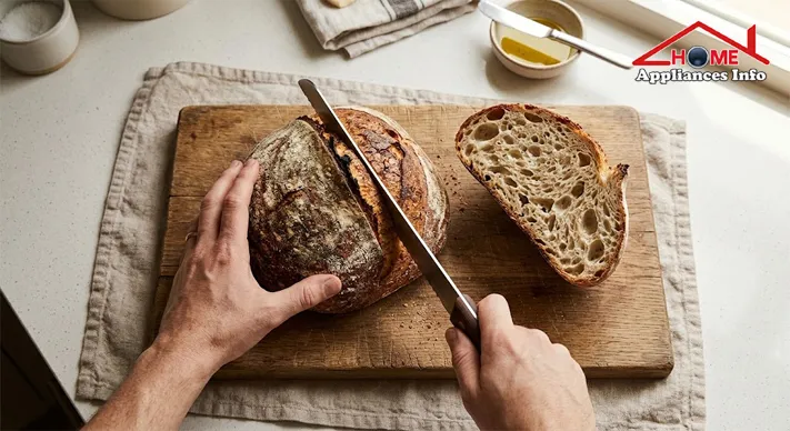 Hands slicing a sourdough boule with a serrated bread knife, the cut half revealing an open crumb
